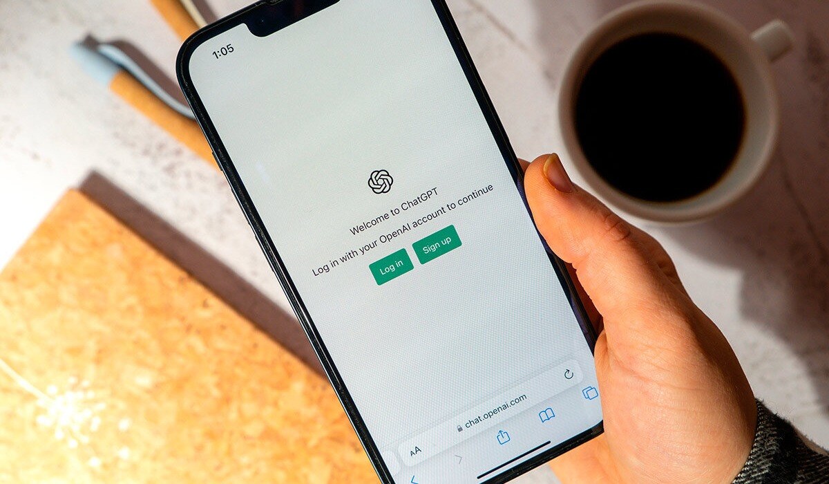 Persona sosteniendo un smartphone con la pantalla de inicio de sesión de ChatGPT, junto a una taza de café sobre una mesa de trabajo.