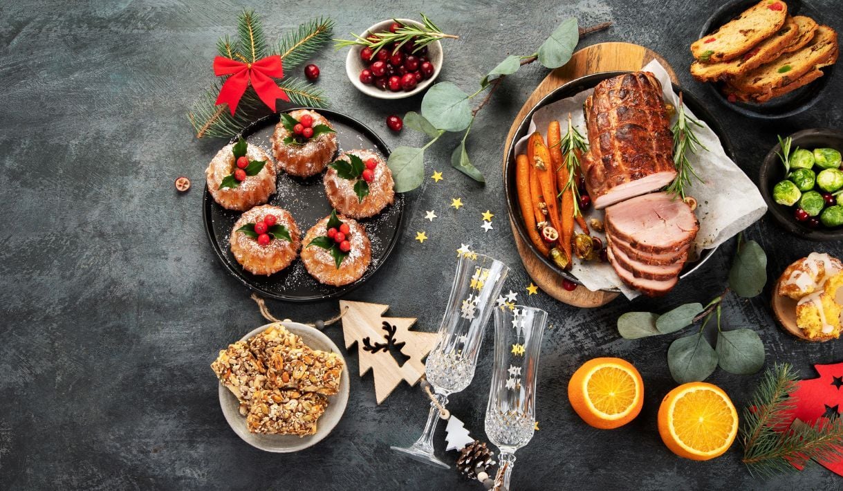 Mesa navideña con platos variados: roscos decorados con frutos rojos, asado de carne con zanahorias, panettone, verduras, copas de brindis y detalles festivos.