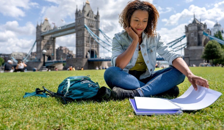 Una joven sentada sobre un césped delante del Puente de la Torre de Londres estudia inglés.