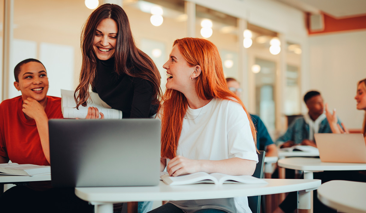 Tres estudiantes universitarias en una sala de estudio, sonriendo y trabajando con portátil abierto