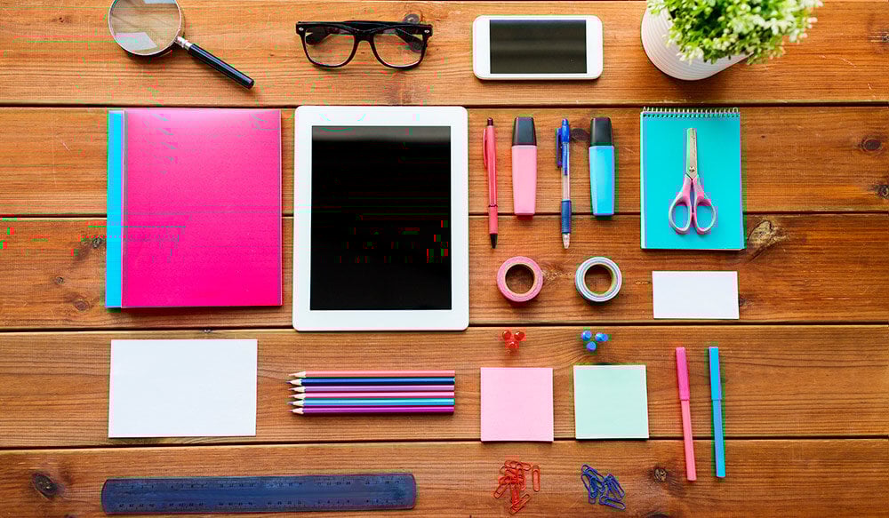Vista superior de una mesa con objetos tradicionalmente asociados a la docencia: una tablet, una lupa, gafas, lapiceros de colores, bolígrafos, tijeras, rollo de celo, clips de colores y notas adhesivas (post-its). La composición refleja herramientas tanto clásicas como modernas para la enseñanza y la organización académica.