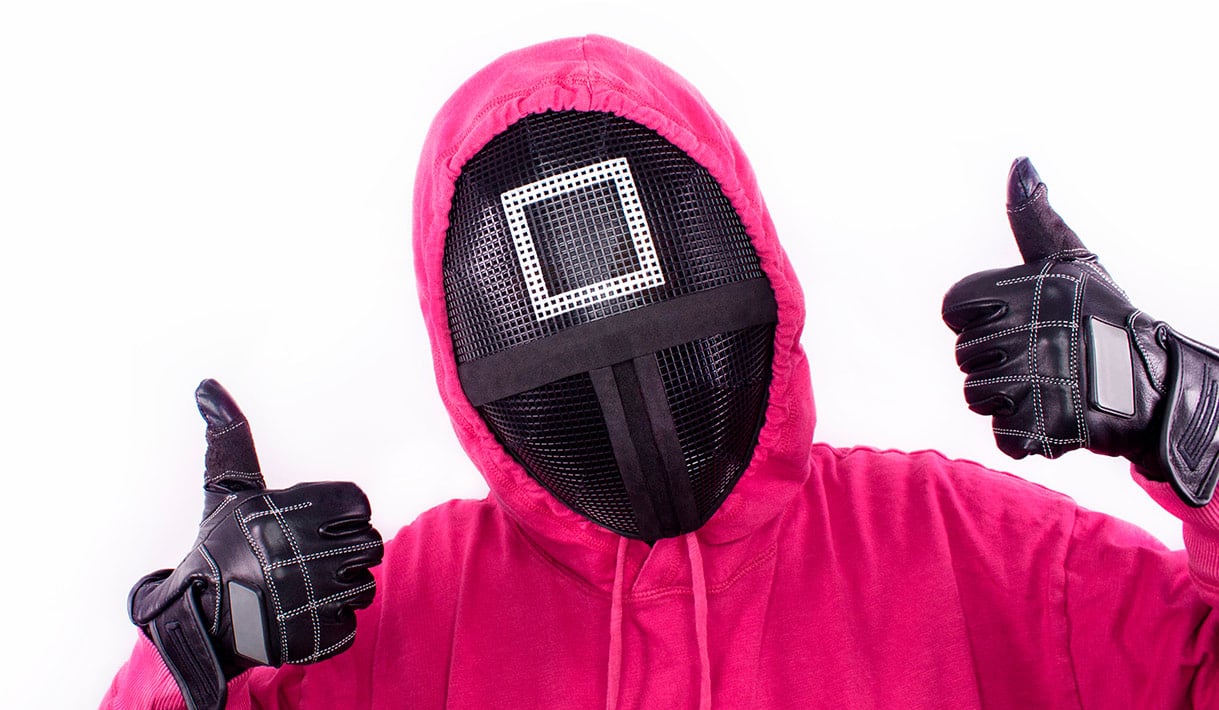 Persona con sudadera roja y máscara negra con símbolo cuadrado, levantando ambos pulgares, inspirada en la serie El juego del calamar.